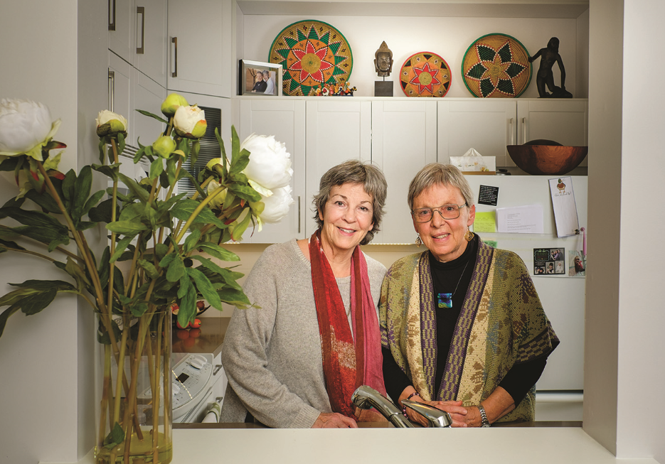 Two smiling older women standing in a kitchenette with a vase of white flowers in the foreground